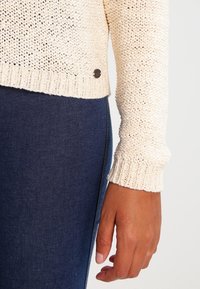 Bras dans un pull en maille crème reposant à côté d'un pantalon en denim bleu foncé sur un fond uni.
