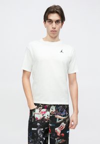 Λευκό βαμβακερό μπλουζάκι με κοντά μανίκια και μαύρο λογότυπο Jordan στο στήθος, σε συνδυασμό με μαύρο παντελόνι που έχει γραφικό με θέμα το μπάσκετ.