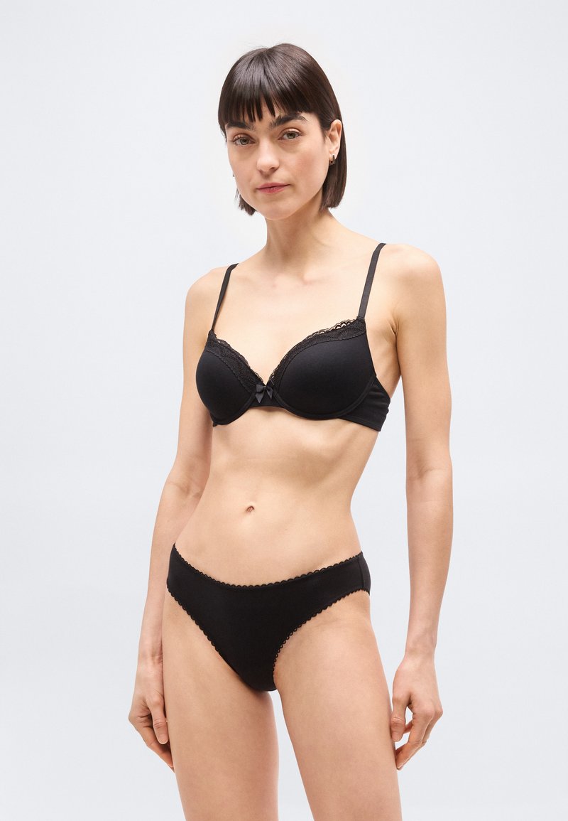 Γυναίκα με κοντά σκούρα μαλλιά, φορώντας ταιριαστό μαύρο σουτιέν και εσώρουχα με δαντέλα, στέκεται μπροστά σε απλό φωτεινό φόντο.