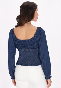 Blusa in denim scuro con maniche lunghe a sbuffo, vita arricciata e scollatura quadrata. Texture liscia con dettagli arricciati alle spalle e all'orlo.