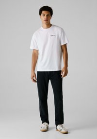 Camiseta de algodón blanca de manga corta, con un pequeño logo impreso. Combinada con pantalones de mezclilla oscuros y zapatillas blancas con suelas de goma.