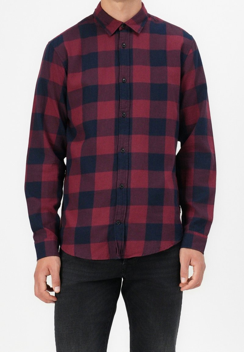 Homme portant une chemise à manches longues à carreaux rouge et bleu foncé, boutonnée, avec un pantalon noir, debout devant un fond blanc uni.