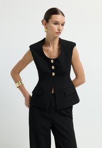 Gilet nero su misura con scollo rotondo, senza maniche, quattro bottoni dorati e due tasche frontali; abbinato a pantaloni neri.