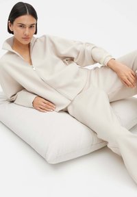 Beige relaxed-fit zip-up jacket with matching pants, featuring a high collar and cuffed sleeves, layered over a soft, neutral cushion.