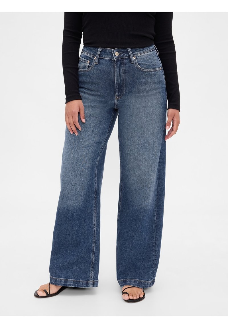 Personne portant un jean bleu taille haute à jambes larges, associé à des sandales noires à lanières et un haut noir à manches longues, debout sur un fond blanc.