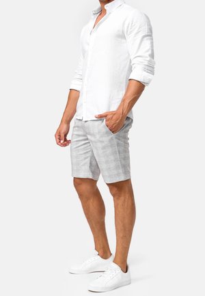 Weißes Leinenhemd mit Knöpfen, kombiniert mit hellgrauen Shorts mit Taschen. Das Modell trägt weiße Sneaker. Einfaches, strukturiertes Design.