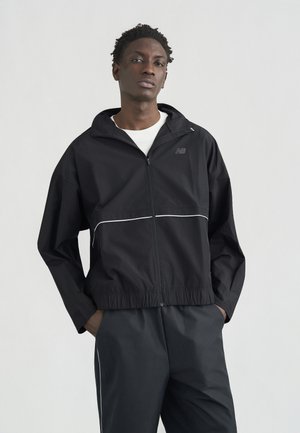 Giovane uomo in piedi con le mani nelle tasche, indossa una giacca nera con zip e pantaloni abbinati con strisce bianche su uno sfondo chiaro uniforme.