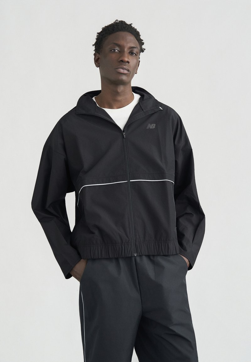 Νεαρός άνδρας στέκεται με τα χέρια στις τσέπες, φορώντας μαύρο μπουφάν με φερμουάρ και αντίστοιχο παντελόνι με λευκές ρίγες σε απλό ανοιχτό φόντο.