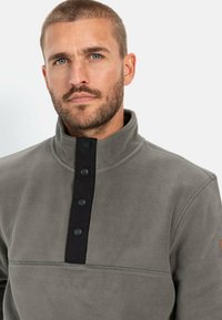 Grå fleecepullov med en sort placket, der har knapper. Høj krave, blød tekstur og kontrastsyningsdetalje.