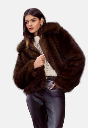 Brown faux fur jacket with a large hood, ribbed texture, and a front zipper, worn over a cream cable-knit sweater and black pants.