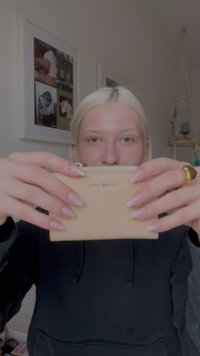 Blondynka trzyma beżową, fakturowaną saszetkę Coccinelle z zamkiem błyskawicznym przed twarzą, ubrana w czarną bluzę z kapturem i złoty pierścionek, w pomieszczeniu z obrazami na ścianie za nią.