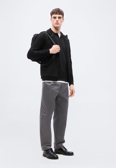 Jeune homme portant une veste noire, un polo noir, un pantalon gris, des chaussures noires, avec un sac à dos noir porté sur une épaule, se tenant devant un fond blanc.