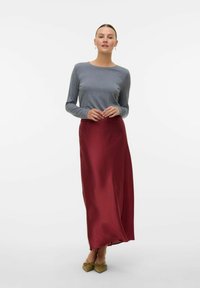 Maglia grigia a maniche lunghe e una gonna in satin borgogna fluida con una texture liscia. La modella indossa scarpe verde oliva, in piedi davanti a uno sfondo neutro.