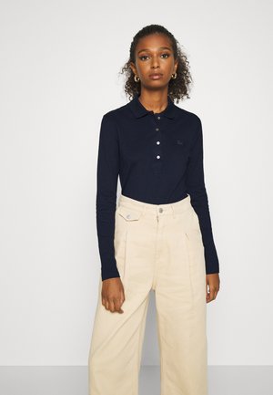 Kvinde med krøllet hår iført navyblå langærmet polo og højtaljede beige vidde bukser, stående mod en ensfarvet lys baggrund.
