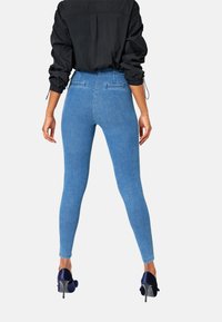Blauwe high-waisted denim leggings met een slim fit en achterzakken, gecombineerd met een zwarte oversized jas en marineblauwe hoge hakken.