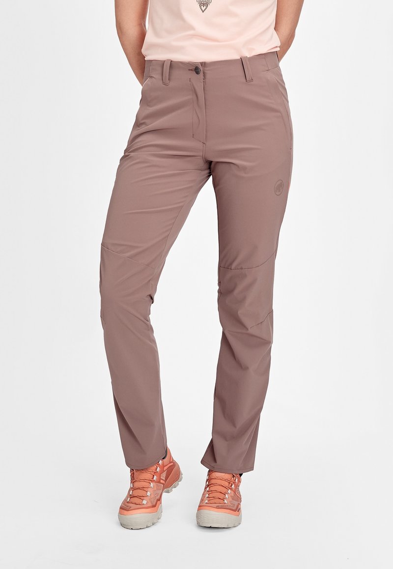 Femme portant un pantalon de randonnée mauve et des chaussures de trail couleur pêche, debout sur un fond blanc uni.