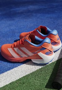 Orange athletic shoes with white stripes, blue accents, textured surface, and a sturdy sole. Features a dynamic design for performance.