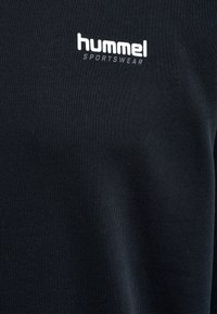 Zbliżenie czarnej tkaniny z białym logo "hummel SPORTSWEAR" nadrukowanym na obszarze klatki piersiowej ubrania.