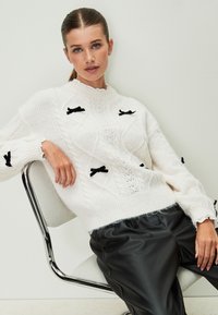 Suéter blanco de punto con patrones de trenzas y detalles de lazos negros; combinado con pantalones de cuero sintético negro; sentado en una silla plateada.