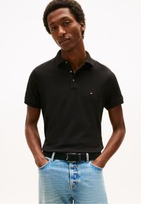 Muž v černé polokošili s krátkým rukávem a světlých modrých džínách s rukama v kapsách na jednobarevném pozadí.