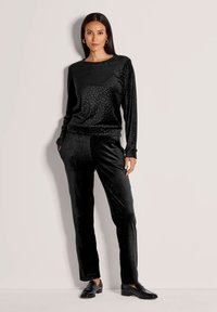 Vrouw in een zwart fluwelen top met lange mouwen met subtiele panterprint en bijpassende zwarte fluwelen broek, staand met handen in haar zakken.