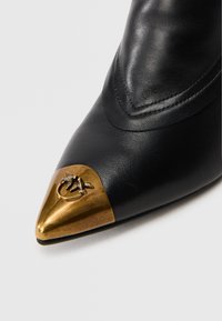 Zapato de cuero negro con punta afilada y una tapa metálica dorada con un motivo de abeja. Textura suave con costuras visibles cerca de la parte superior.