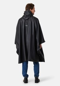 Svart vattentät poncho med huva, försedda med tryckknappar på sidorna, slät textur och en logotyp på baksidan. Bärs över blå jeans.