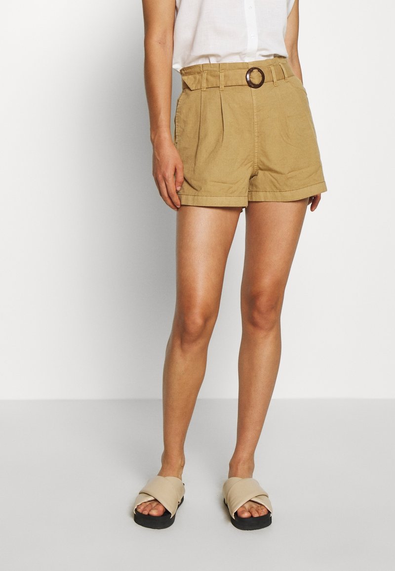 Femme portant un short plissé beige taille haute avec une ceinture, un haut blanc et des sandales beige à lanières croisées, debout sur un fond uni.