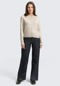 Beżowy kardigan z długim rękawem, okrągłym dekoltem i guzikami z przodu, połączony z ciemnymi, szerokimi spodniami dżinsowymi oraz czarnymi butami. Gładka faktura.