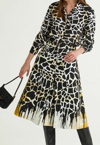 Woman wearing black and white giraffe print buttoned dress with yellow accents, holding a small black purse and accessorized with a red ring.