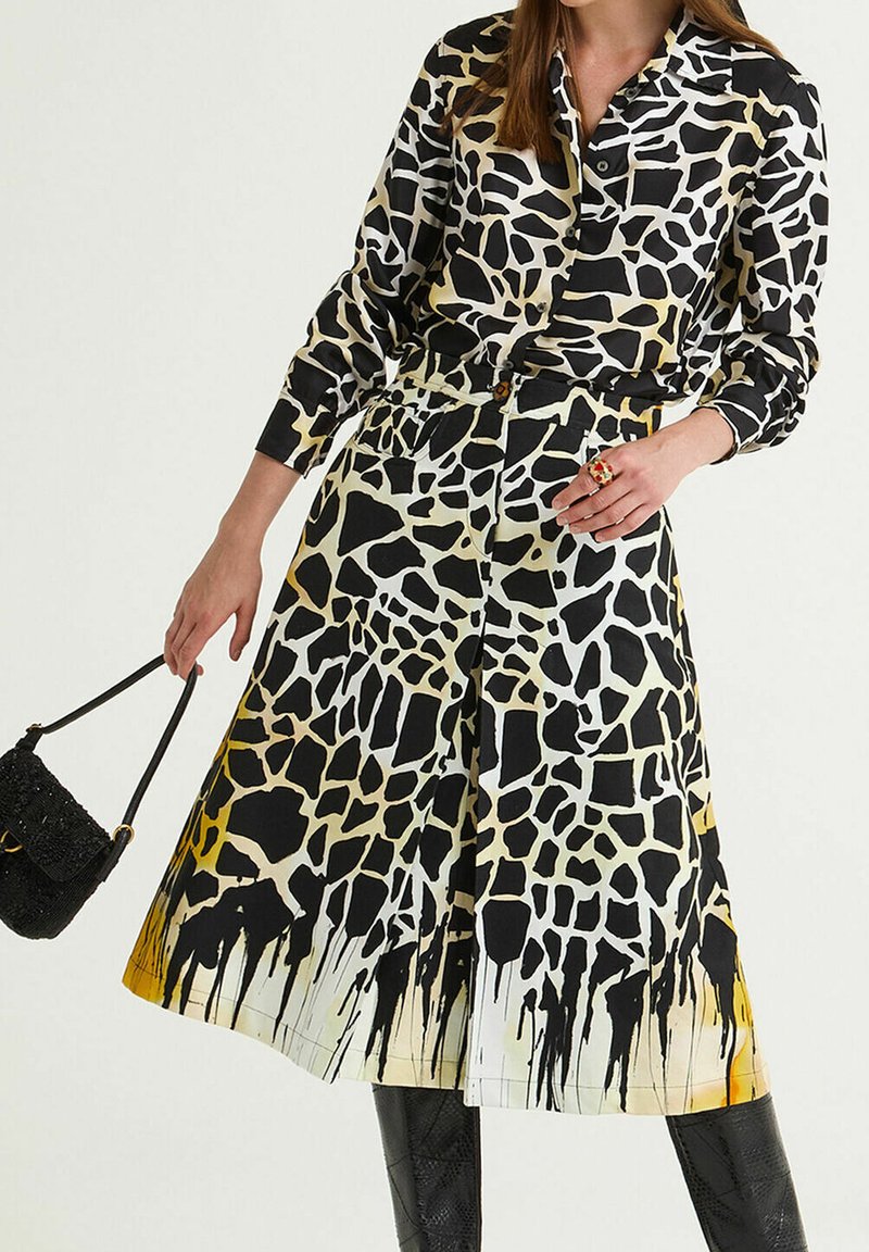 Woman wearing black and white giraffe print buttoned dress with yellow accents, holding a small black purse and accessorized with a red ring.
