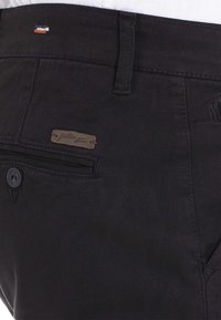 Un pantalon en tissu noir présente une petite étiquette de logo marron, une poche avant avec fermeture à bouton, et un design épuré avec un minimalisme dans les détails.