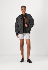 Juodas bomber tipo švarkas su oranžine vidine puse, derinamas su baltais raštuotais šortais ir juodais slip-on batais. Trumpi plaukai, stovintis vertikaliai.