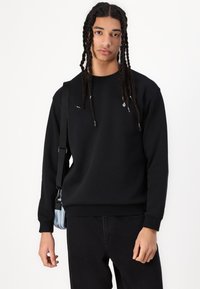 Schwarzer Pullover aus weichem Stoff, mit Rundhalsausschnitt, gerippten Bündchen und kleinem Logodetail. Kombiniert mit schwarzen Hosen und einer Tasche.