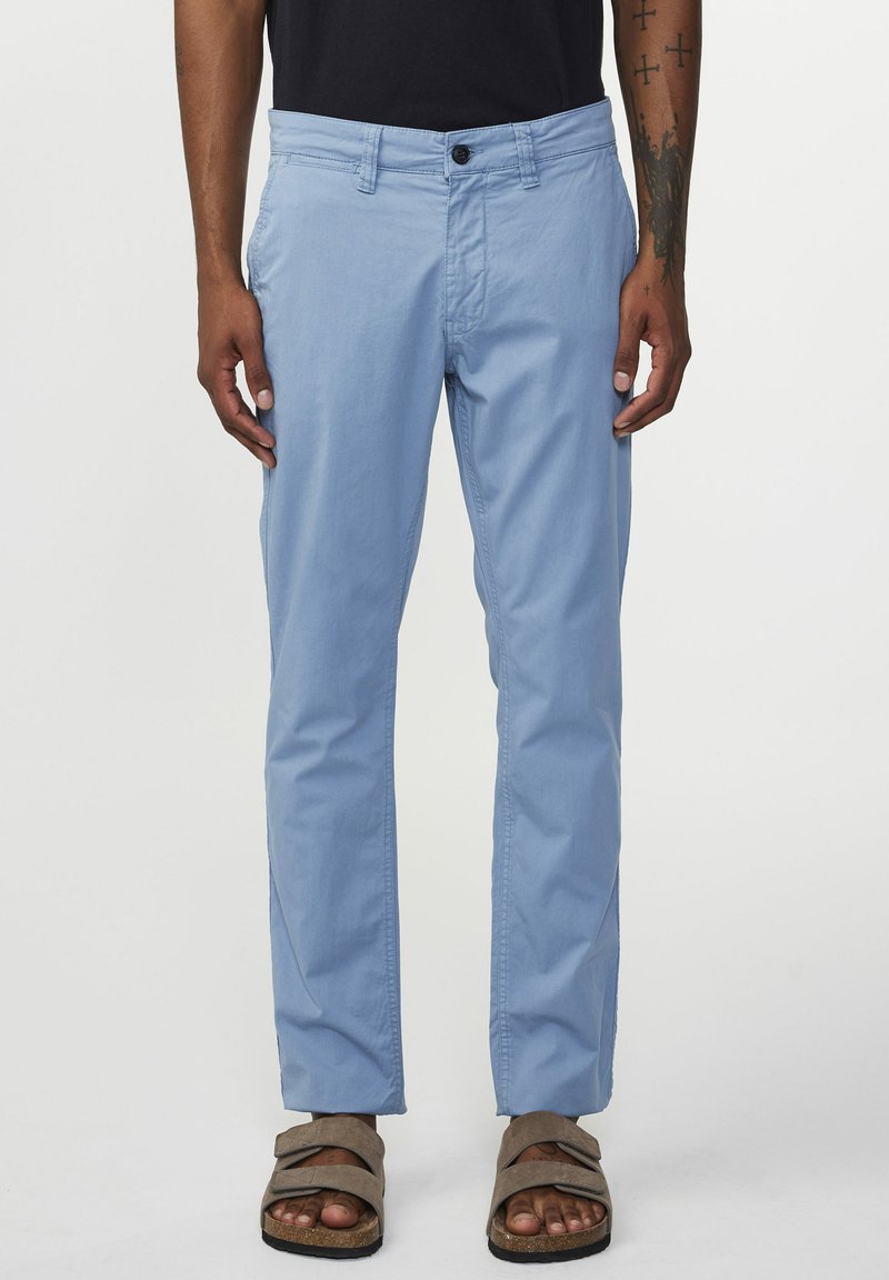 Pantaloni di cotone azzurri chiari con chiusura a bottone, taglio dritto e tasche laterali, abbinati a sandali marroni. Dettagli di cucitura minimal.