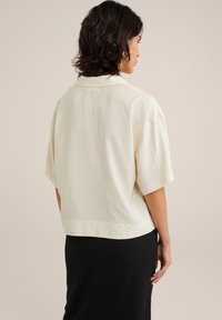 Bluză cu mânecă scurtă, de culoare crem, dintr-un material ușor; are guler și o croială ușor scurtă, de tip boxy, cu o textură netedă.