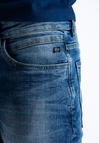 Niebieskie dżinsy o wyblakłym odcieniu, z podwójnym ściegiem, pięcioma kieszeniami i małą metką marki na tylnej kieszeni.