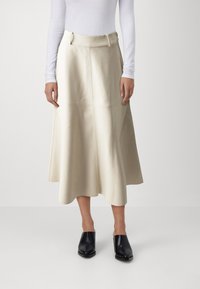 Jupe midi en cuir couleur crème avec un design évasé, des coutures visibles et une ceinture avec une quincaillerie minimaliste. Associée avec des mules à talons carrés noirs.