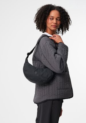Schwarze Nylon-Schultertasche mit runder Form, zwei Knopfakzenten und einem verstellbaren Tragegurt, getragen von einer Person in einer gepolsterten grauen Jacke.