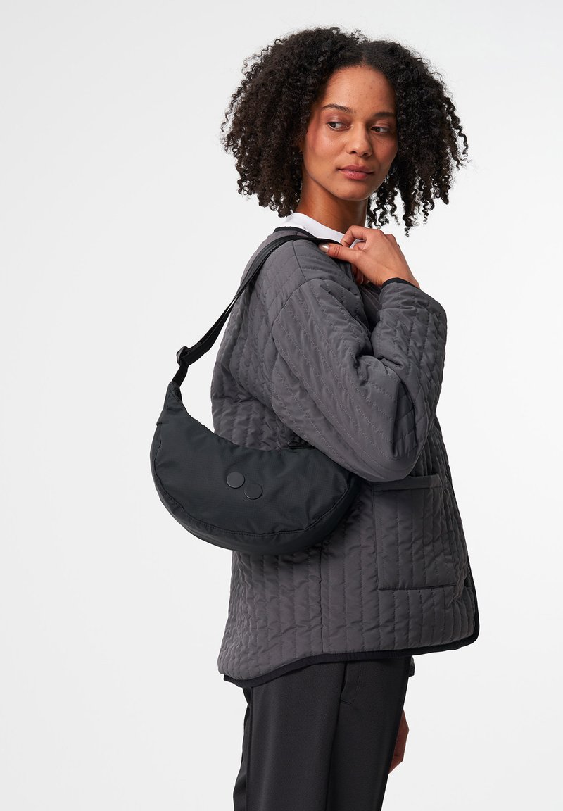 Schwarze Nylon-Schultertasche mit runder Form, zwei Knopfakzenten und einem verstellbaren Tragegurt, getragen von einer Person in einer gepolsterten grauen Jacke.