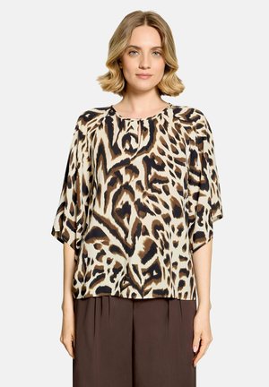 Kvinde med kort blondt hår iført en løs leopardprint-top og mørkebrune højhalsbukser, der står mod en ensfarvet baggrund.