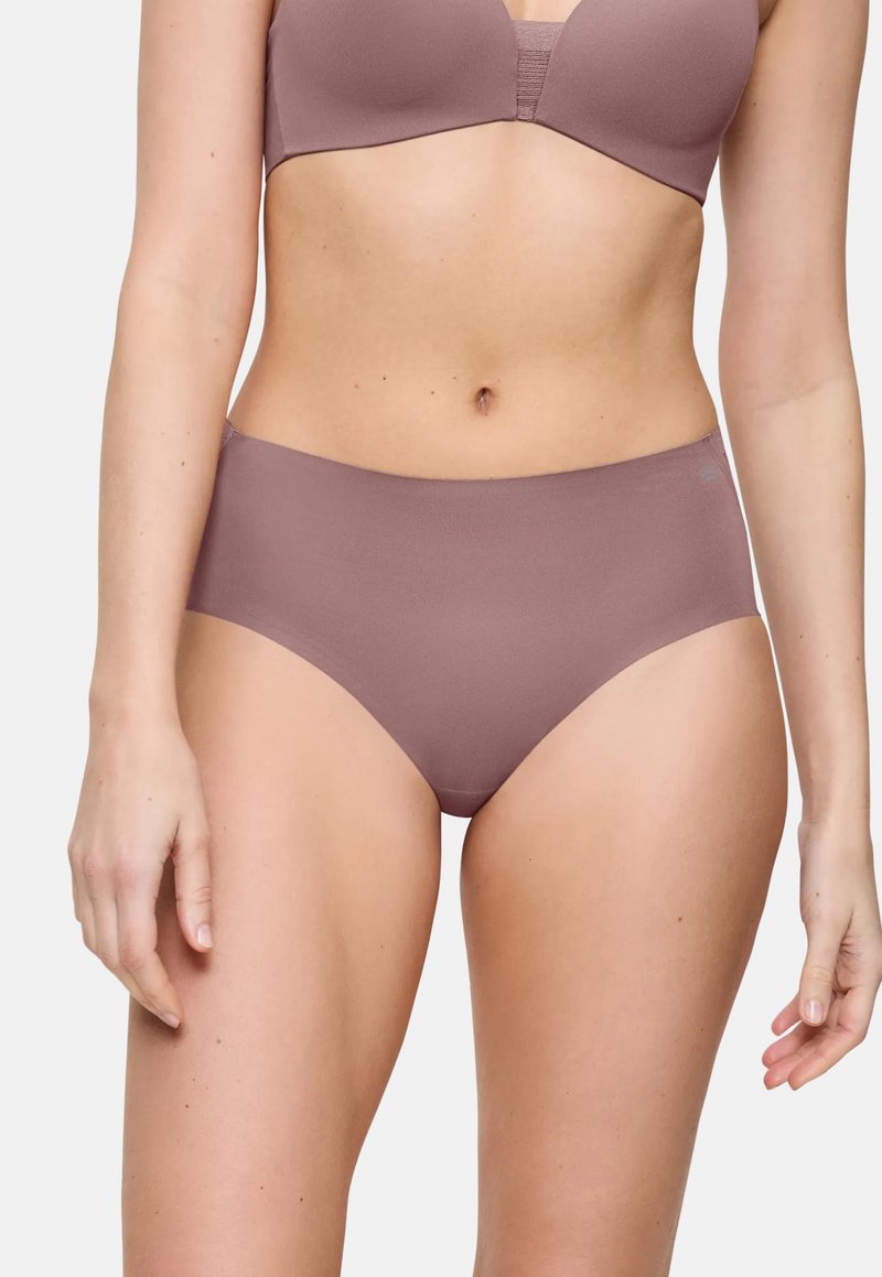 Femeie îmbrăcată într-un sutien mauve fără cusături și chiloți asortați cu talie înaltă, stând în fața unui fundal deschis simplu.