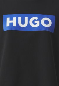 Čierne tričko s výrazným, obdĺžnikovým modrým logom, na ktorom je v strede hrubými bielymi písmenami napísané "HUGO".