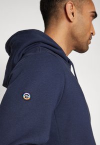 Sweat à capuche bleu marine en tissu doux, doté d'une poche kangourou et d'un patch logo rond sur l'épaule gauche.