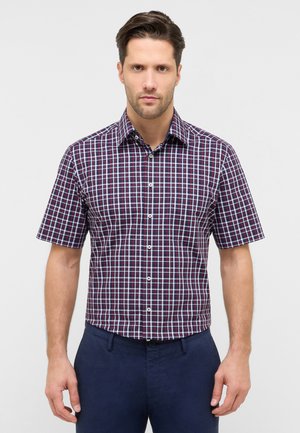 Uomo che indossa una camicia aderente a maniche corte con motivo a quadri rosso, bianco e blu e pantaloni blu scuro davanti a uno sfondo semplice.