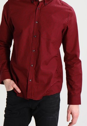 Man draagt een rood-zwart klein geruit overhemd met lange mouwen en knopen, gecombineerd met een zwarte broek, met één hand in de zak en de andere ontspannen naast zich.