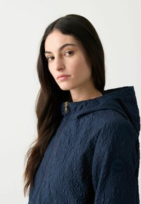 Navyblauwe textuurjack met een capuchon, ritsluiting en gouden hardwaredetails. Voorzien van een logo patch op de linker mouw.