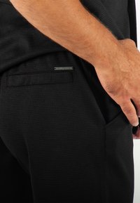 Pantalons noirs texturés avec une coupe ajustée. Présentent une poche latérale et une petite étiquette métallique avec "HOLLYGHOST" près de la couture de la poche.