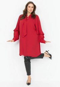 Sieviete pozē uz vienas kājas, uzvilkusi sarkanu garu piedurkņu blūzi ar loku, melnus ādas bikses un melnas augstas papēža kurpes uz balta fona.