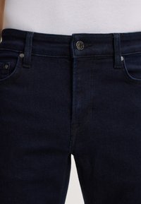 Jeans in denim blu scuro con chiusura a cerniera, tasche frontali e bottone metallico. Il tessuto ha una finitura liscia e leggermente testurizzata.
